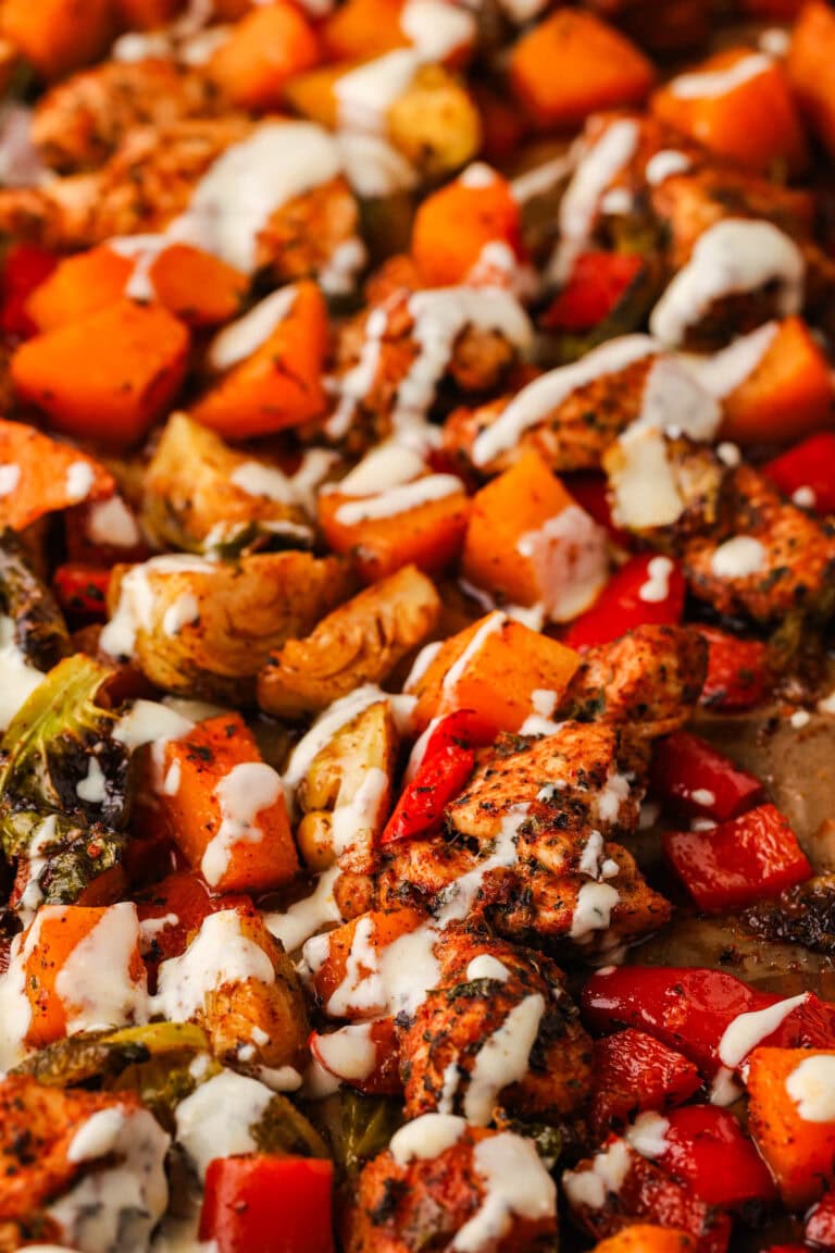 A close-up of a roasted sheet pan chicken breast meal with seasoned red bell peppers, Brussels sprouts, sweet potatoes, and a drizzle of creamy white sauce.