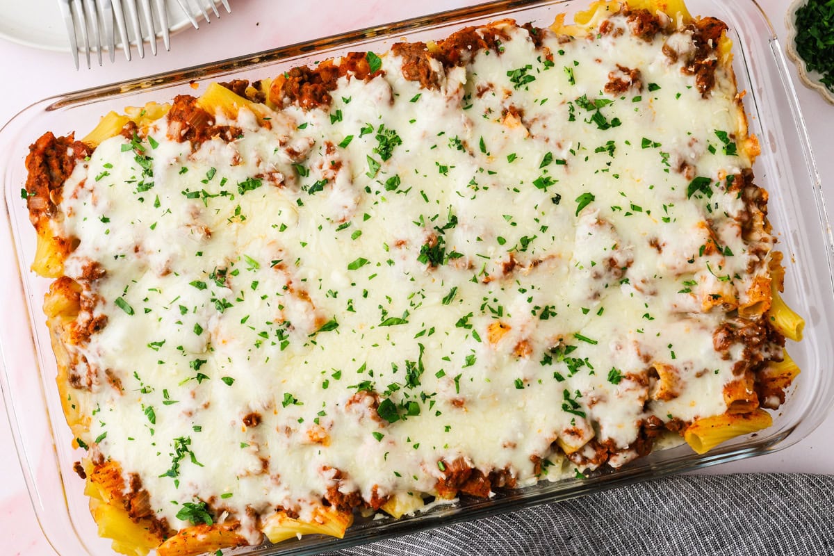A glass baking dish filled with baked ziti topped with melted cheese, browned meat sauce, and sprinkled with chopped parsley—a dish that proves not all meals are fails of pasta. Plates and a gray striped towel are visible nearby.