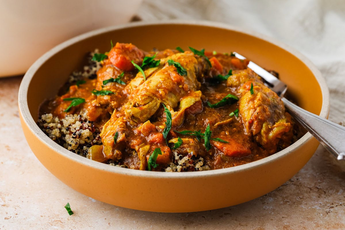 A shallow brown bowl filled with chicken curry inspired by chicken wing stew, garnished with fresh herbs and served over a bed of quinoa. A fork rests in the bowl, against a softly lit, neutral-toned background.
