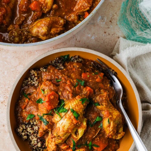 A bowl filled with chicken wing stew in a rich tomato sauce served over quinoa, garnished with chopped parsley. A fork rests in the bowl, and the stew pot is seen above with more chicken and sauce.