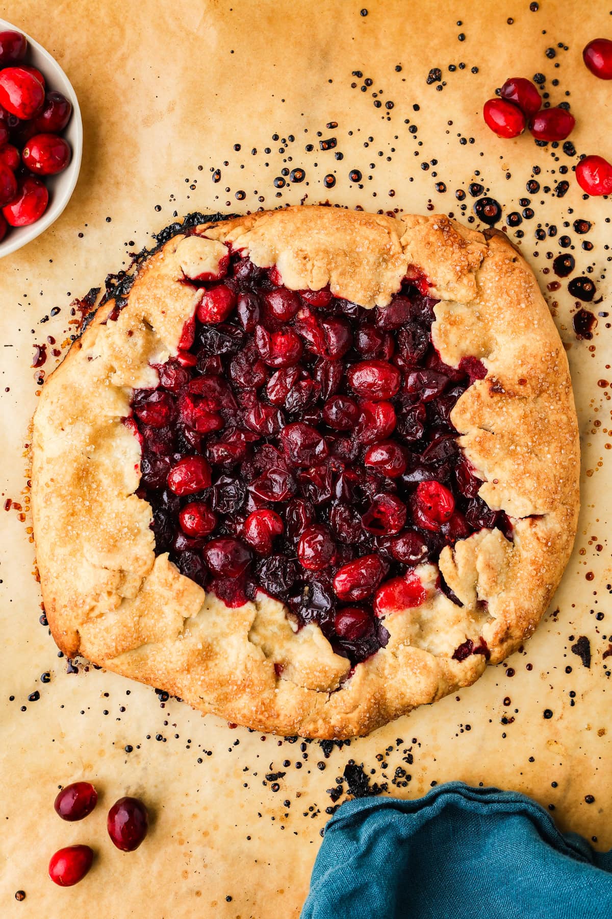 A rustic cranberry galette with a golden, flaky crust is displayed on parchment paper. Fresh cranberries are scattered around, and part of a blue cloth napkin is visible in the corner.