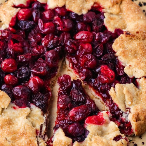 A rustic cranberry galette with a golden, flaky crust and vibrant red cranberry filling. One slice has been cut and slightly separated from the rest of the tart, showing the juicy berries inside.