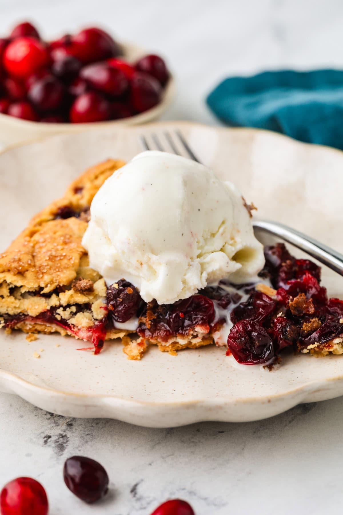 A slice of cherry pie topped with vanilla ice cream sits on a plate with a fork. In the background, a small bowl of fresh cherries and a cranberry galette rest beside a blue napkin.