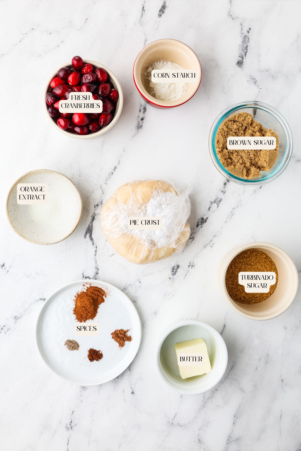 Seven bowls and plates on a white surface display ingredients for a cranberry galette: fresh cranberries, corn starch, brown sugar, turbinado sugar, butter, orange extract, spices, and a wrapped pie crust in the center.