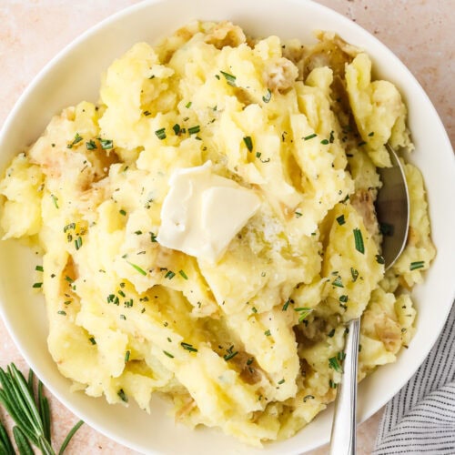 A bowl of mashed potatoes topped with a pat of butter and chopped herbs, with a spoon resting in the bowl. A sprig of rosemary and a striped napkin are beside the bowl on a light surface.