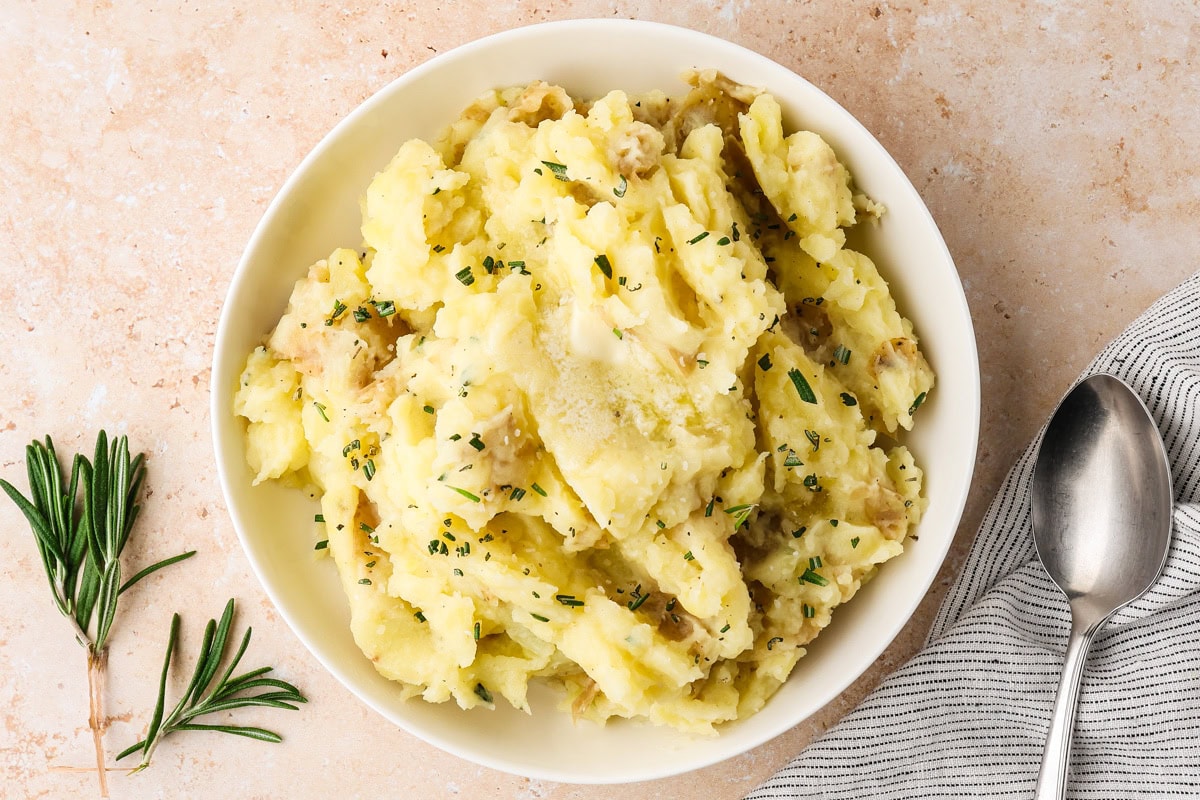 A bowl of mashed potatoes garnished with chopped herbs, placed on a light-colored surface next to a sprig of rosemary, a striped napkin, and a spoon.