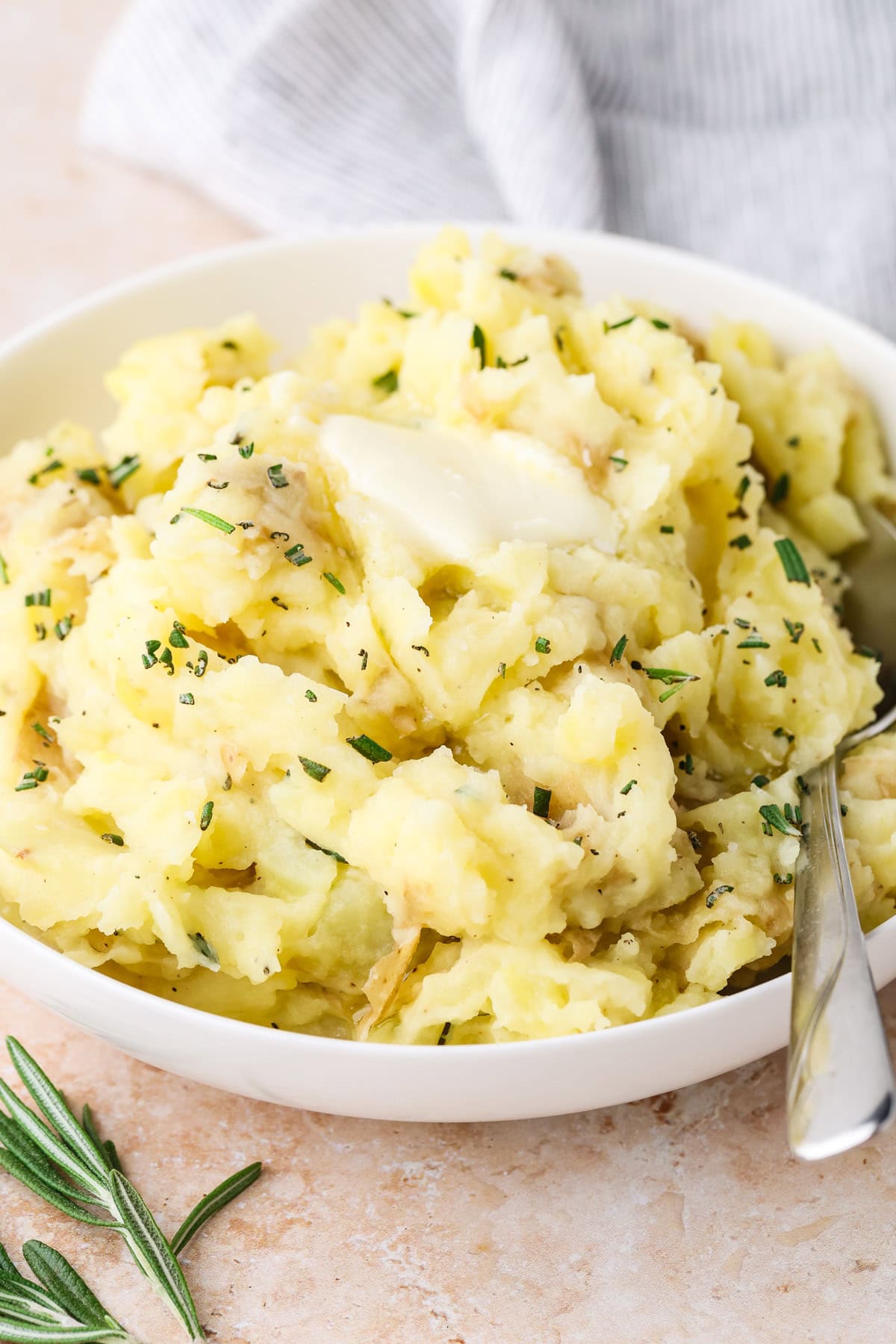 A bowl of creamy mashed potatoes topped with a pat of melting butter and sprinkled with chopped herbs, with a silver spoon beside the potatoes. A sprig of rosemary and a striped cloth are nearby.