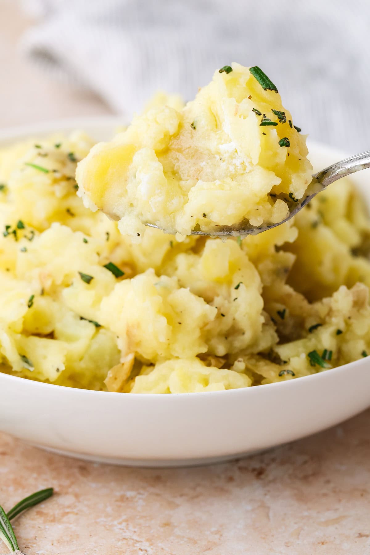 A spoonful of mashed potatoes with herbs is held above a white bowl filled with more mashed potatoes, garnished with chopped green herbs.