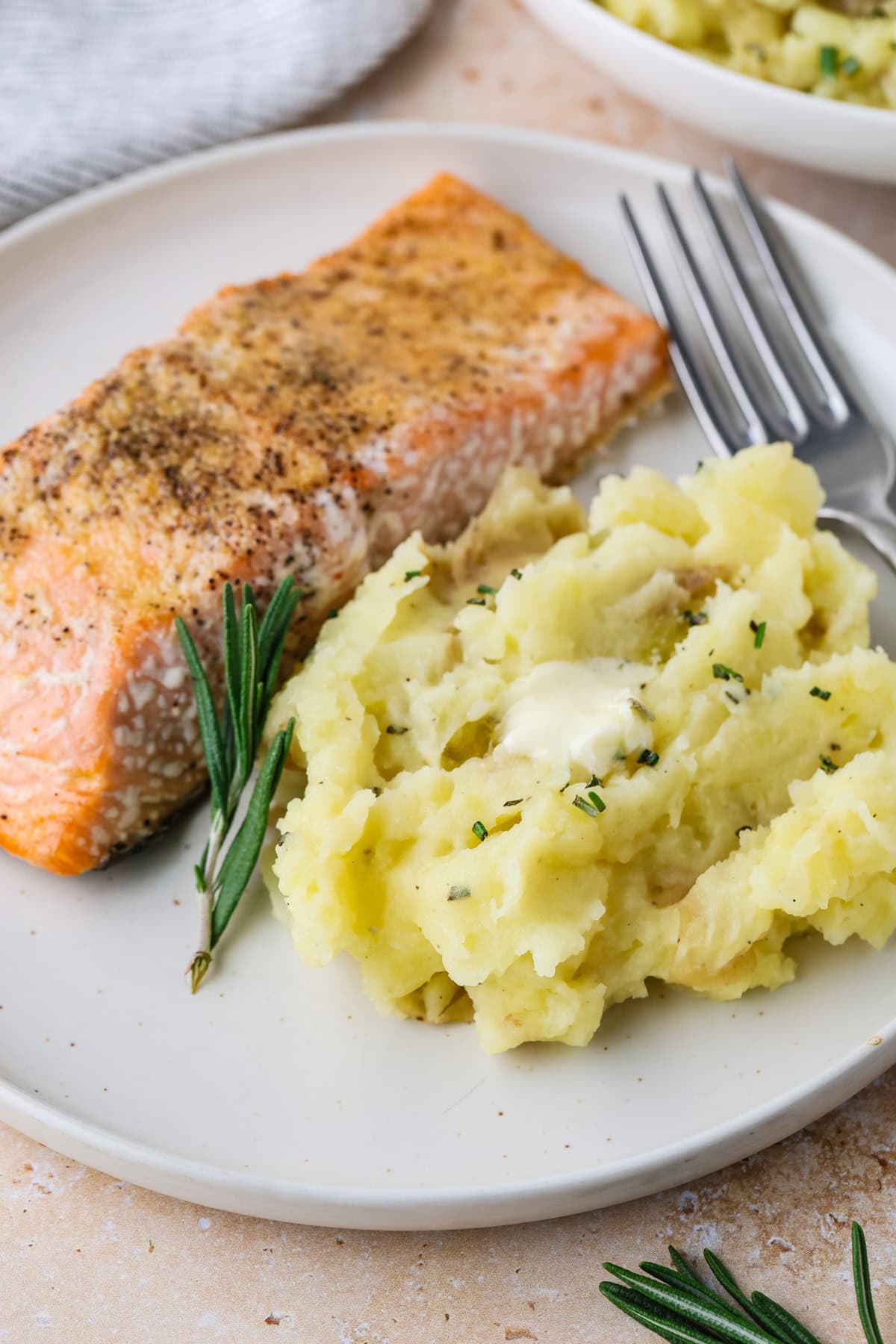 A plate with a seasoned salmon fillet and a serving of mashed potatoes topped with butter, garnished with fresh rosemary. A fork rests on the plate.