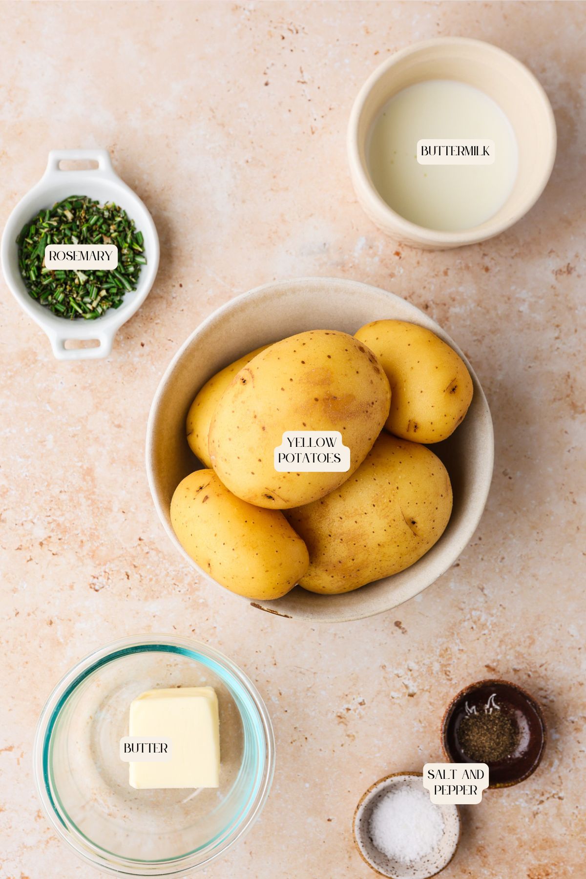 Top-down view of ingredients for mashed potatoes with buttermilk: a bowl of yellow potatoes, buttermilk, chopped rosemary, a chunk of butter, and salt and pepper.