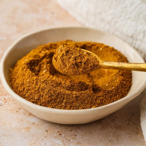 A bowl filled with ground spice, with a golden spoon resting on top and holding a scoop of the spice, sits on a light-colored surface next to a white textured cloth.