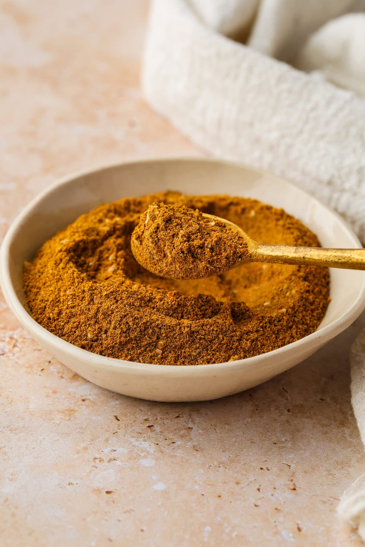 A bowl filled with ras el hanout spice mix with a golden spoon resting on top and holding a scoop of the spice, sits on a light-colored surface next to a white textured cloth.