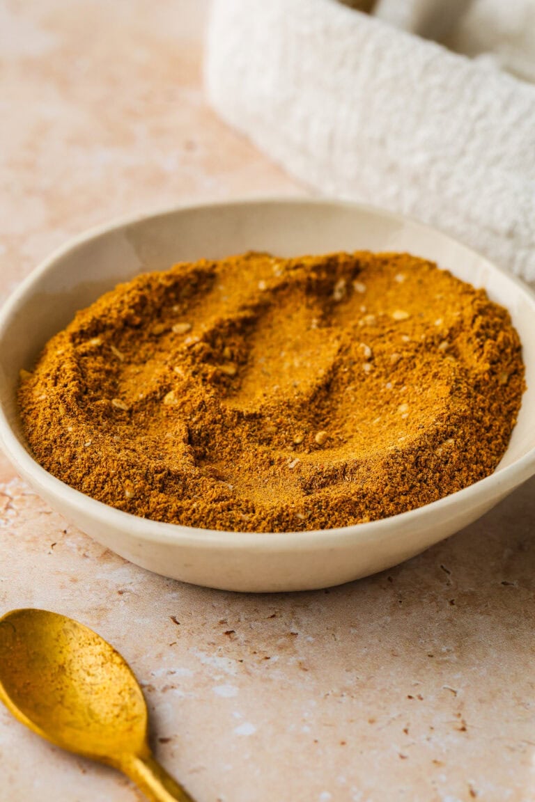 A shallow beige bowl filled with a finely ground, golden-brown spice mix sits on a light stone surface. A small gold spoon with spice residue rests nearby. A textured white cloth is in the background.