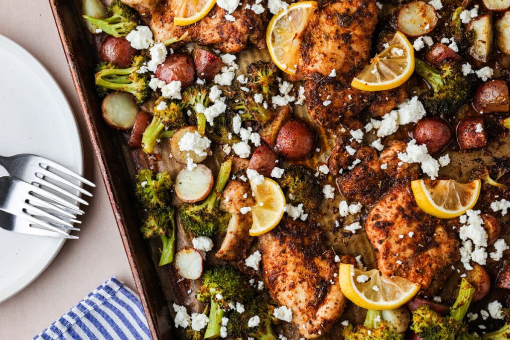 A sheet pan with seasoned baked chicken, broccoli, red potatoes, lemon slices, and crumbled feta cheese. A striped napkin, plate, and forks are on the left side.