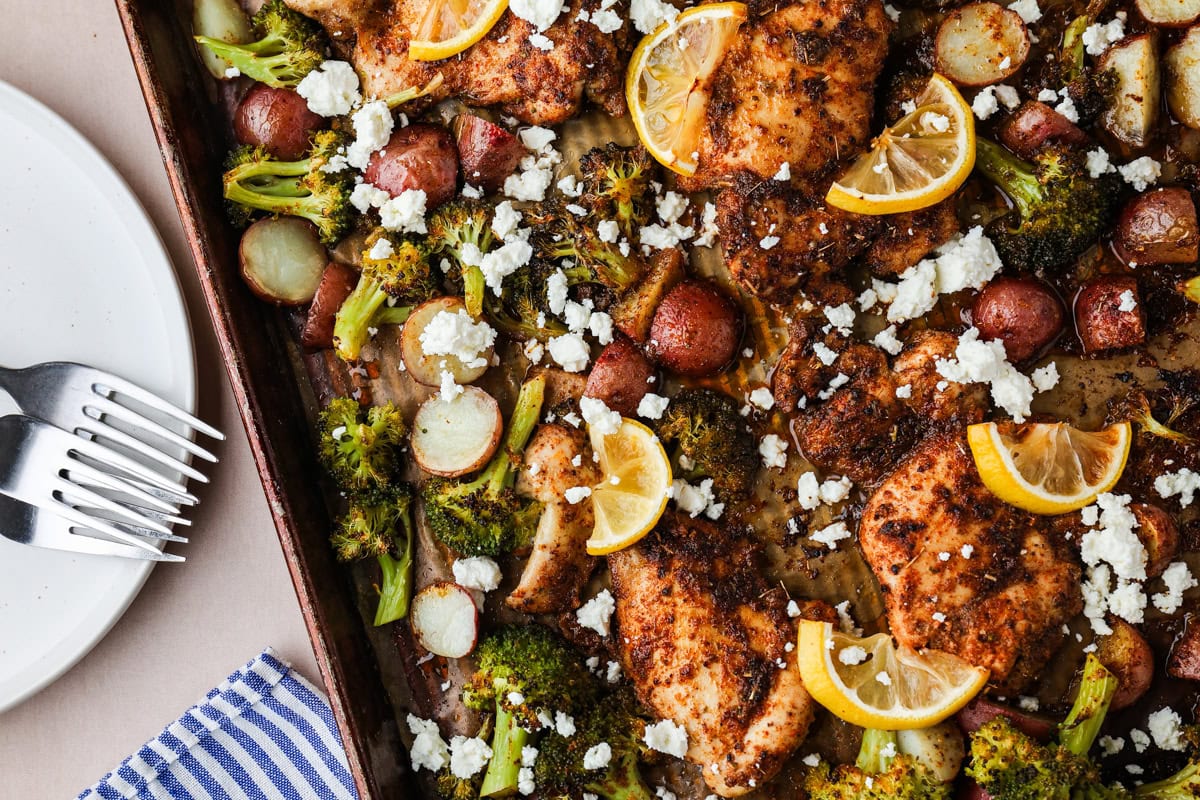 A sheet pan with seasoned baked chicken, broccoli, red potatoes, lemon slices, and crumbled feta cheese. A striped napkin, plate, and forks are on the left side.