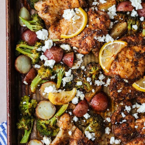 A sheet pan filled with roasted chicken thighs, sliced red potatoes, broccoli florets, lemon slices, and crumbled feta cheese. The ingredients are evenly spread and golden brown.