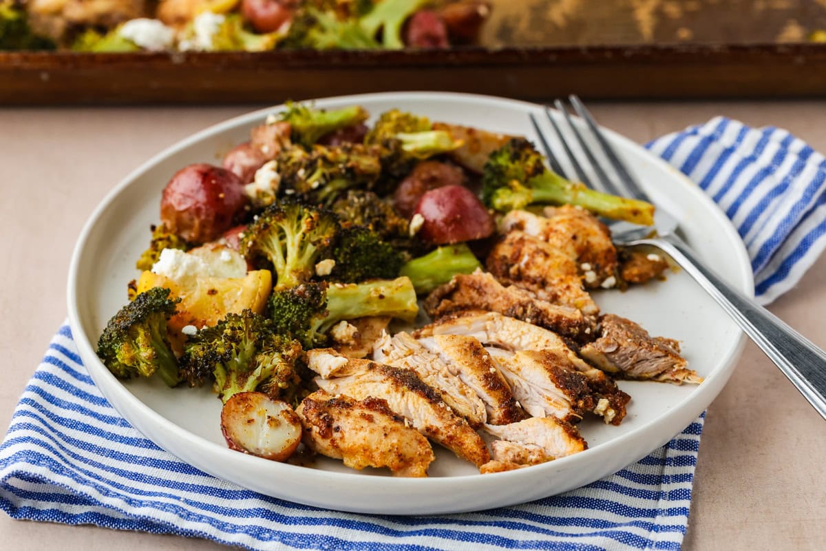A plate with sliced roasted chicken, broccoli, red potatoes, and crumbled cheese, set on a blue and white striped napkin with a fork.