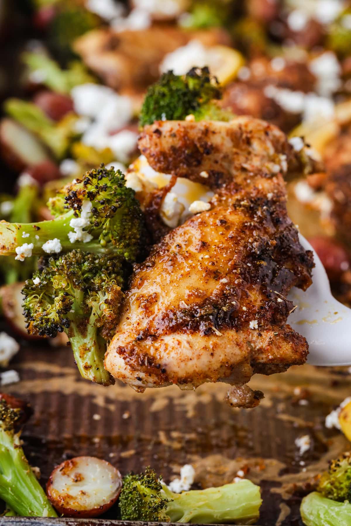 A close-up of a cooked, seasoned chicken thigh with roasted broccoli and crumbled cheese, served on a spatula over a sheet pan with more vegetables and chicken in the background.