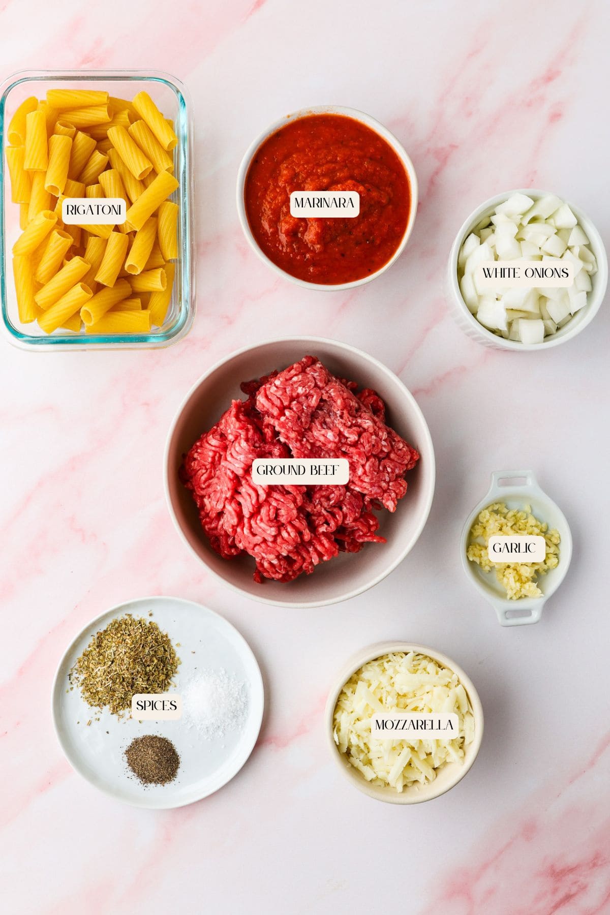 A flat lay of ingredients for a pasta dish, including uncooked rigatoni, marinara sauce, chopped white onions, minced garlic, ground beef, shredded mozzarella cheese, and assorted spices on a pink background—no fails of pasta here!.
