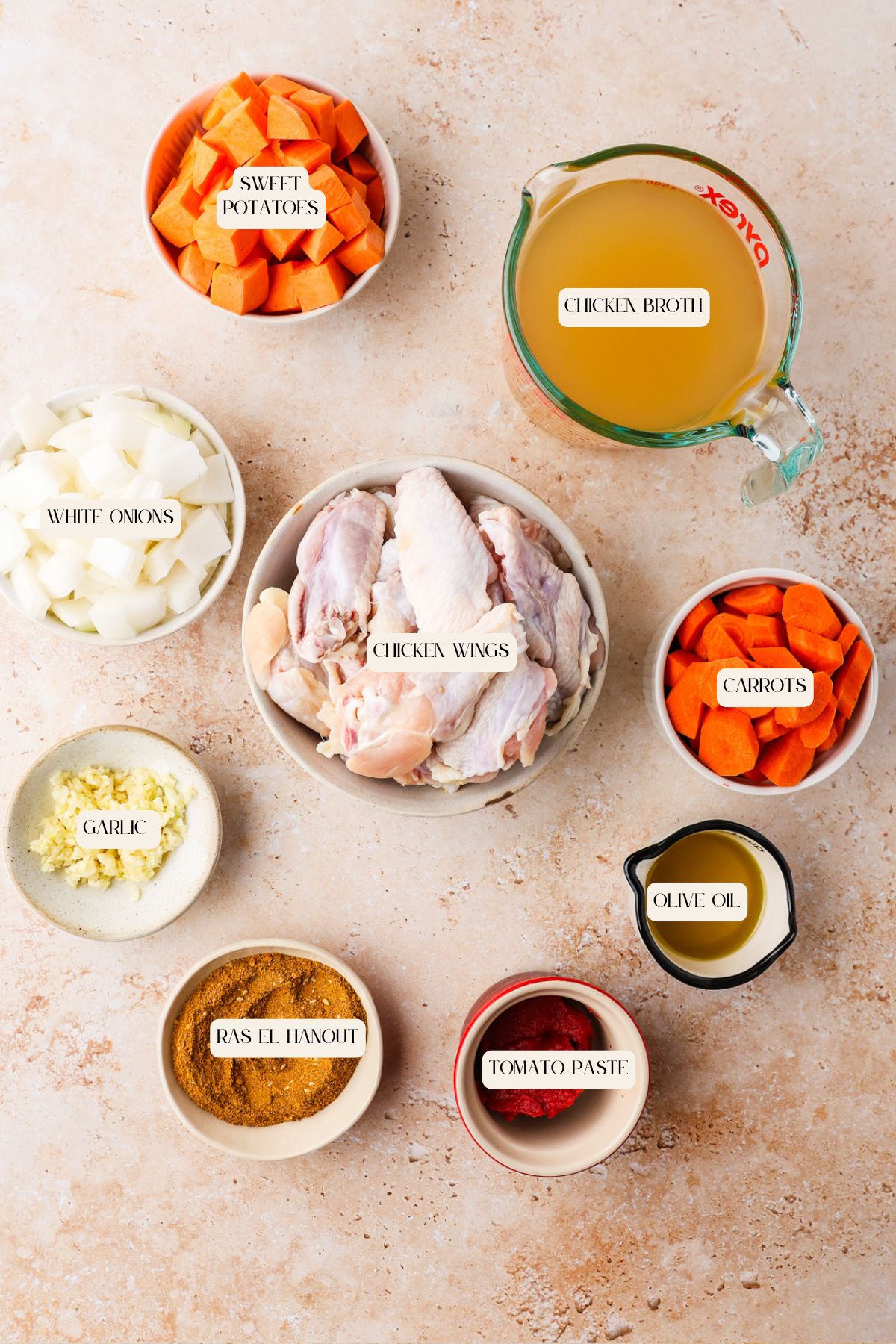 Top-down view of labeled ingredients for chicken wing stew: chopped sweet potatoes, chicken broth, white onions, raw chicken wings, chopped carrots, olive oil, tomato paste, ras el hanout spice, and minced garlic in bowls.