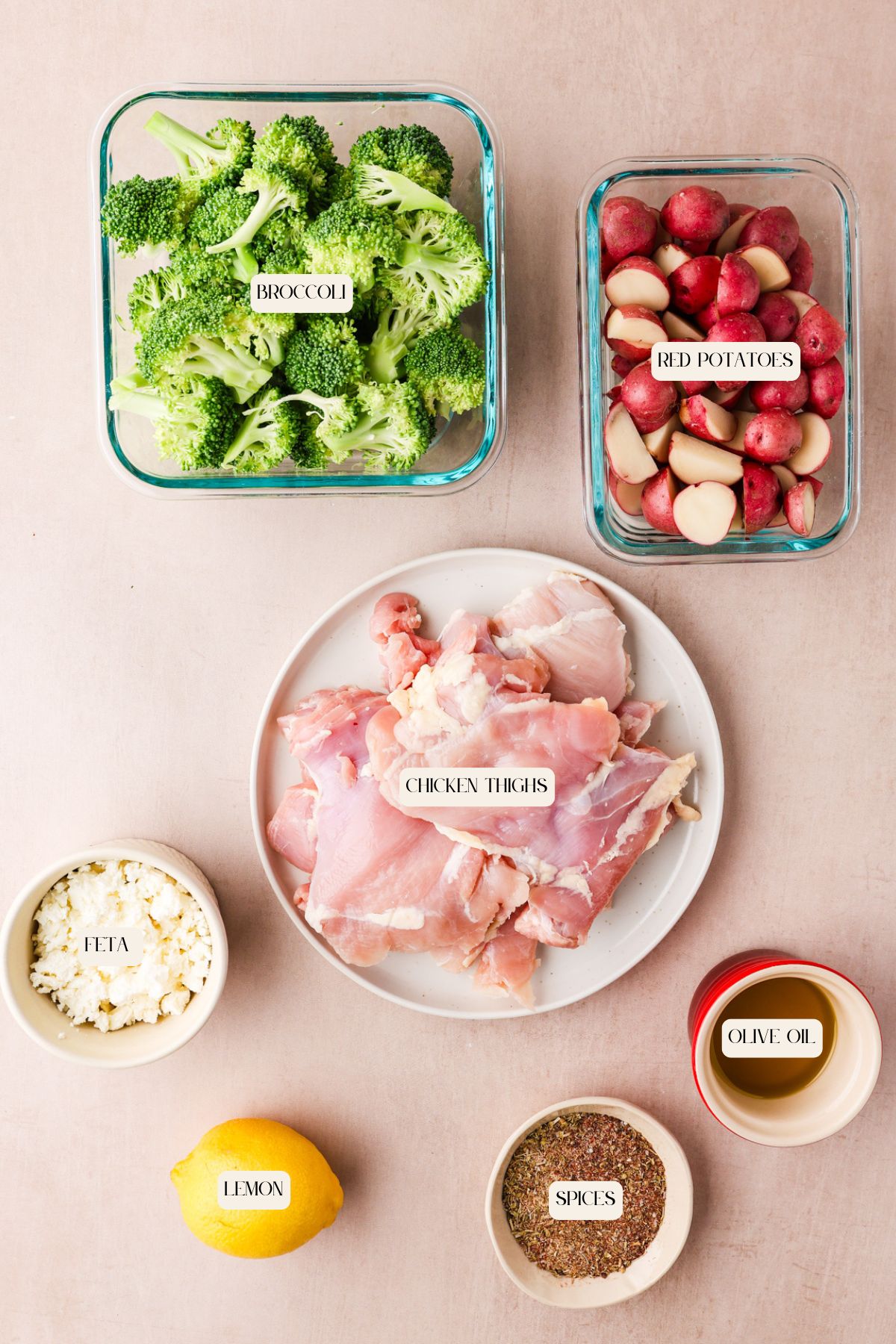 A flat lay of ingredients in separate bowls: chopped broccoli, halved red potatoes, raw chicken thighs, crumbled feta cheese, olive oil, a whole lemon, and a bowl of mixed spices. Each container is labeled.