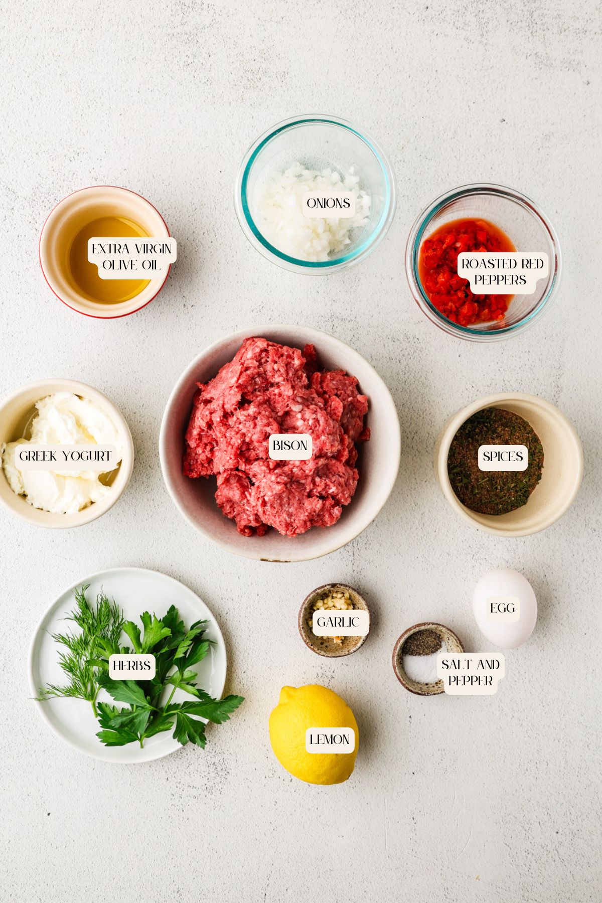 A flat lay of ingredients for bison burgers in bowls labeled: extra virgin olive oil, onions, roasted red peppers, Greek yogurt, ground bison, spices, egg, garlic, salt and pepper, fresh herbs, and a whole lemon on a white surface.