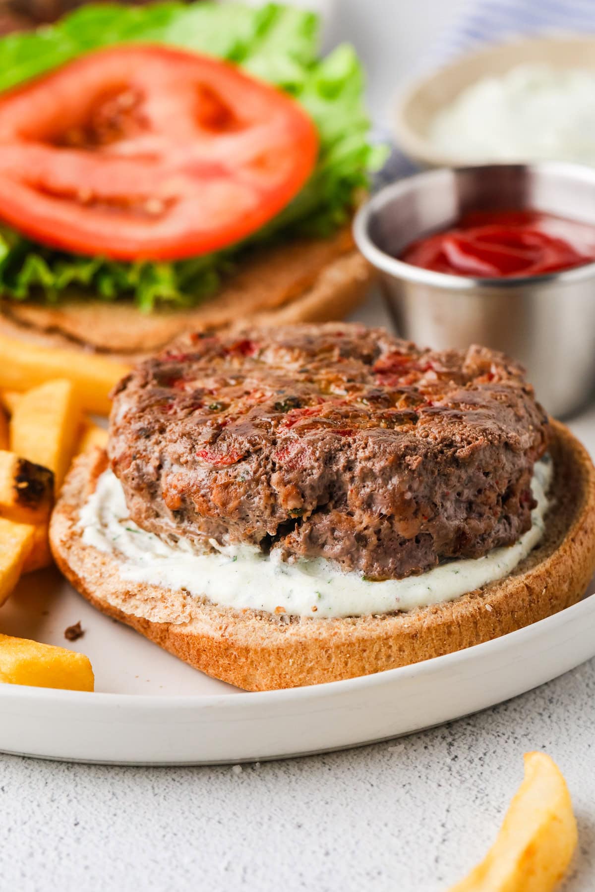 An open-faced bison burger with a beef patty and white sauce on a bun, served with lettuce, tomato slices, French fries, and small cups of ketchup and white dipping sauce on a white plate.