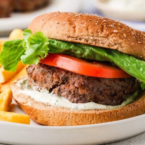 A close-up of a veggie burger in a whole wheat bun, similar in style to classic bison burgers, topped with lettuce, tomato slices, and a spread, served on a white plate with a side of French fries.
