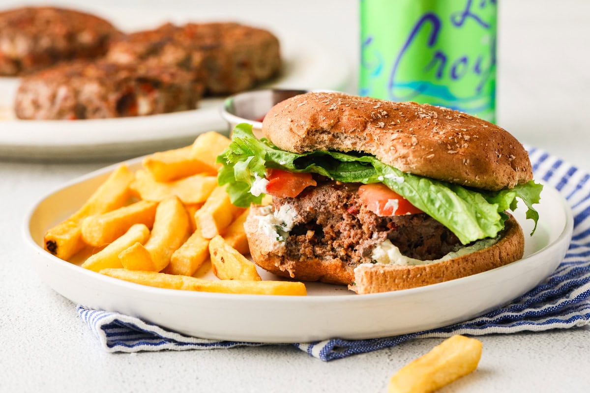 A plate with a partially eaten burger in a whole wheat bun, filled with lettuce and tomato, next to a serving of French fries. A can of La Croix sparkling water and more burgers on a plate are in the background.