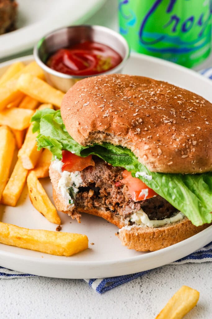A plate with a partially eaten bison burger topped with lettuce and tomato, served with crispy french fries. A small container of ketchup and a can of sparkling water are in the background.