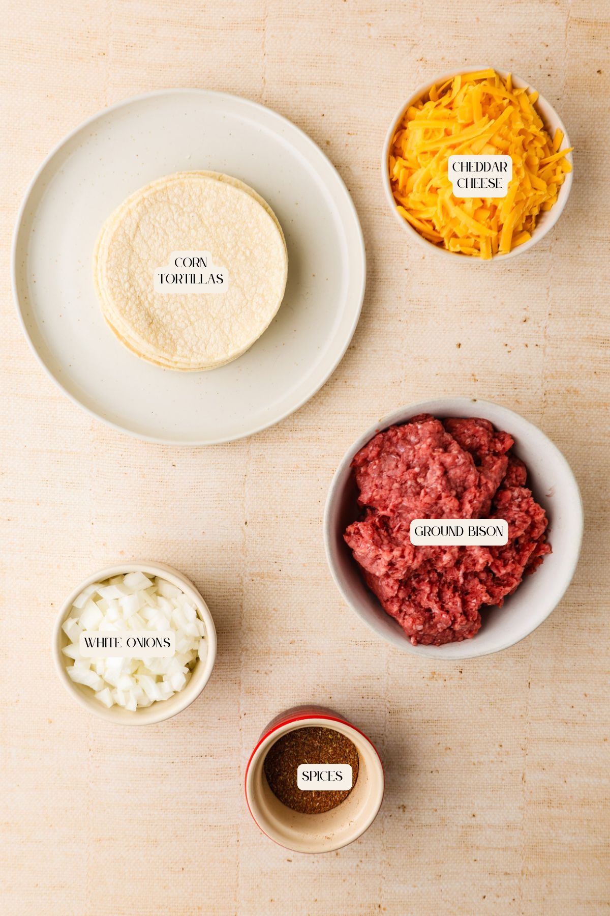 Five bowls and plates containing ingredients for tacos are arranged on a light surface: corn tortillas, shredded cheddar cheese, ground bison, chopped white onions, and a blend of spices.