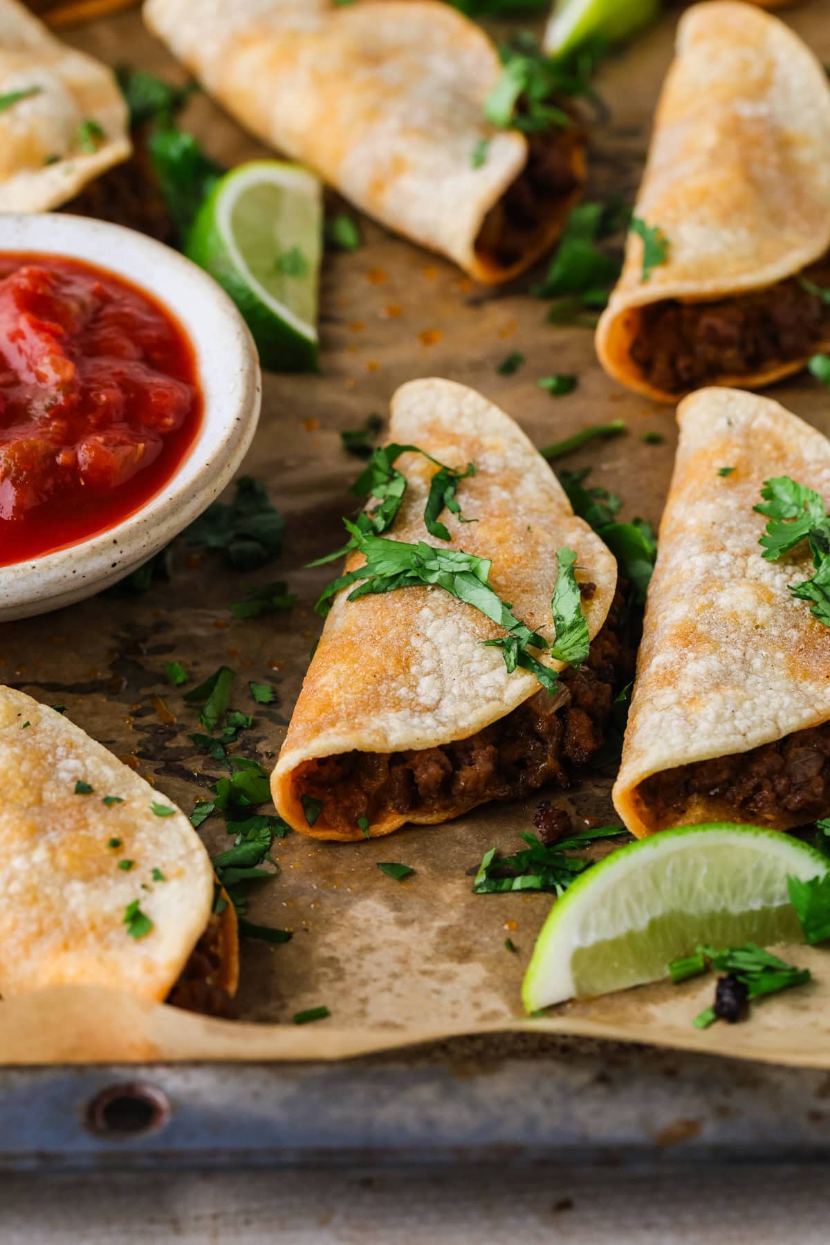 Tacos filled with seasoned ground beef, topped with fresh cilantro, arranged on parchment paper with lime wedges and a bowl of salsa on the side.