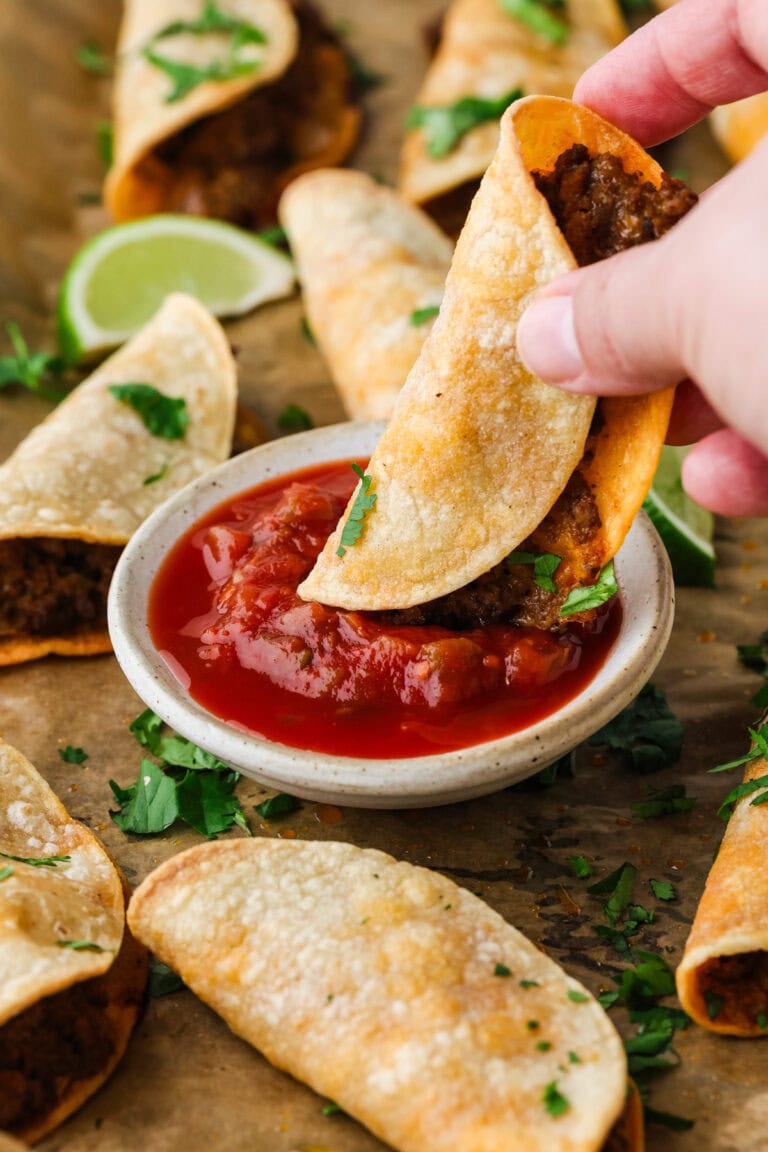 A hand dips a crispy taco filled with meat into a bowl of red salsa, surrounded by more tacos, fresh cilantro, and lime wedges on a brown surface.