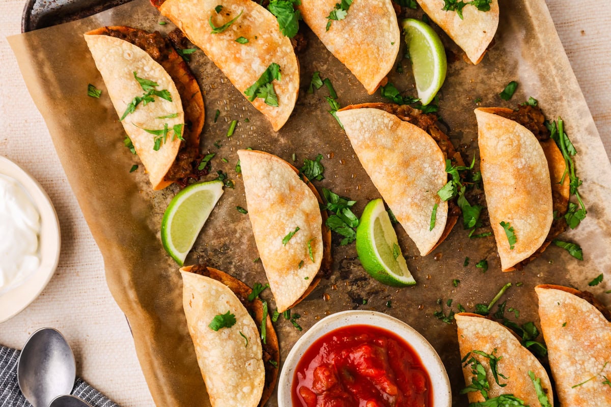 A baking tray with crispy folded tacos filled with a dark filling, garnished with chopped cilantro and lime wedges. A bowl of salsa and a small dish of sour cream are beside the tray.