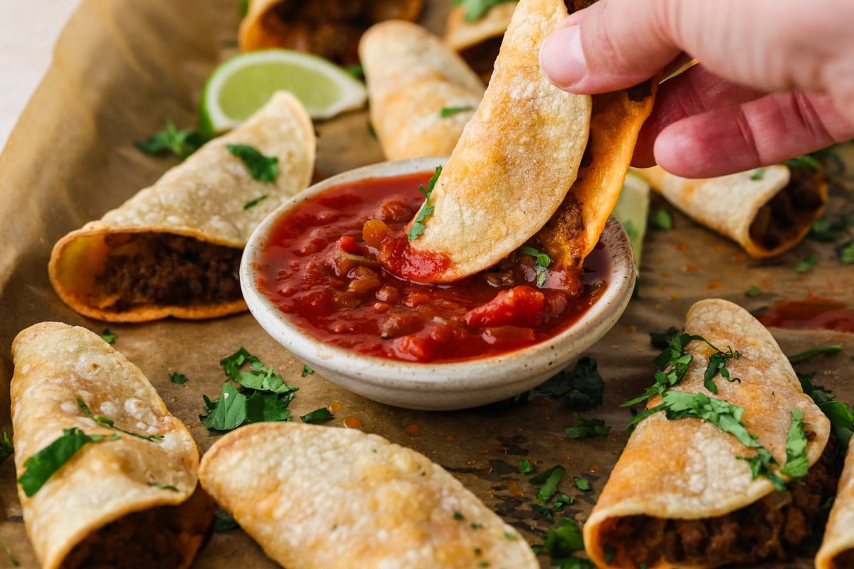 A hand dips a crispy taquito filled with meat into a bowl of red salsa, surrounded by more taquitos garnished with chopped cilantro on parchment paper. A lime wedge is in the background.