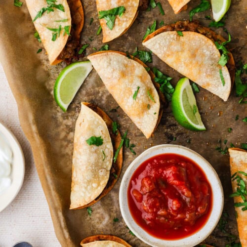 A baking sheet with crispy folded tacos garnished with chopped cilantro, lime wedges, and a small bowl of red salsa. A plate of sour cream and two metal spoons are nearby.