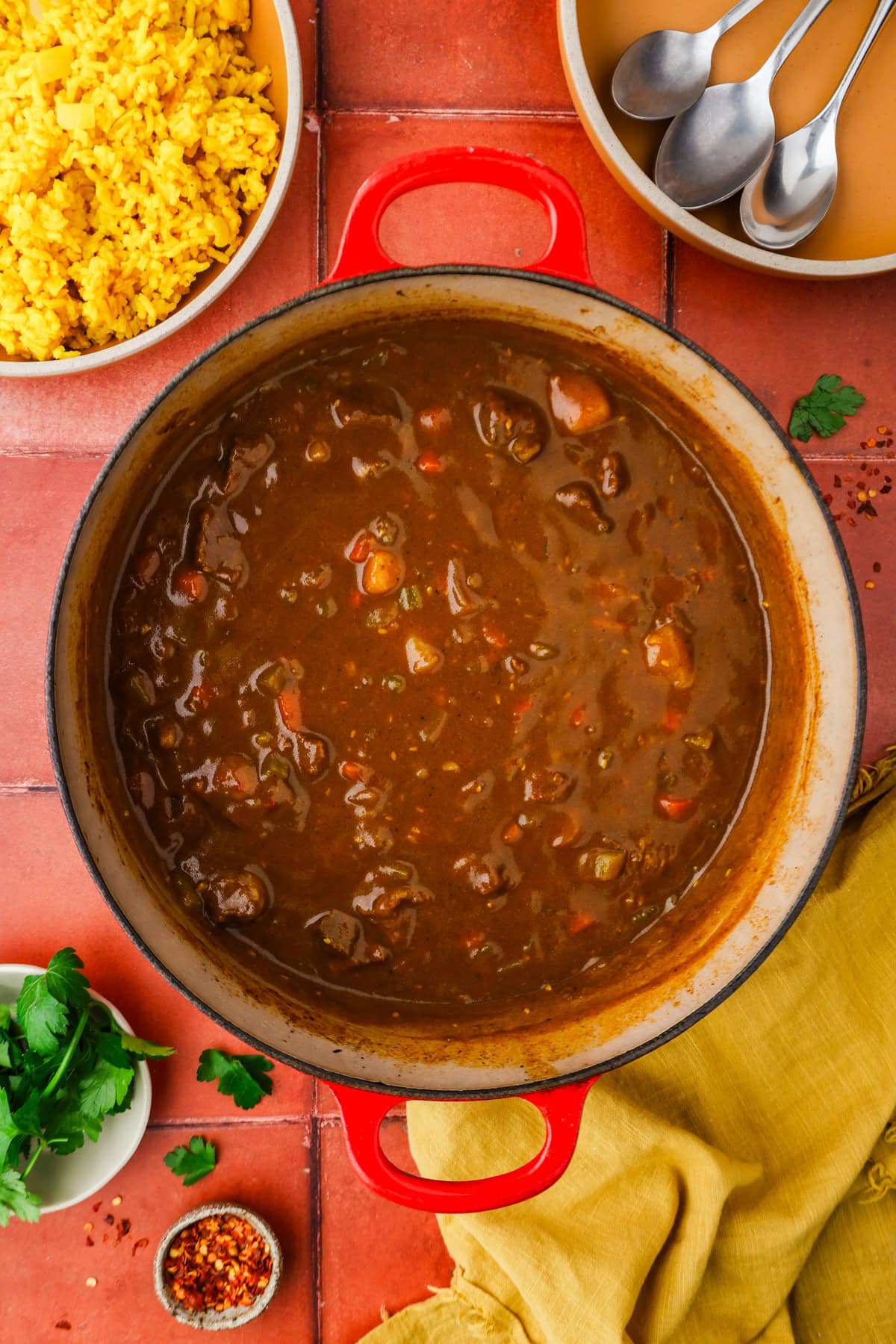 A large red pot filled with dark brown stew containing chunks of meat and vegetables sits on a red tile surface, surrounded by a bowl of yellow rice, a small bowl of cilantro, red pepper flakes, spoons, and a yellow cloth.