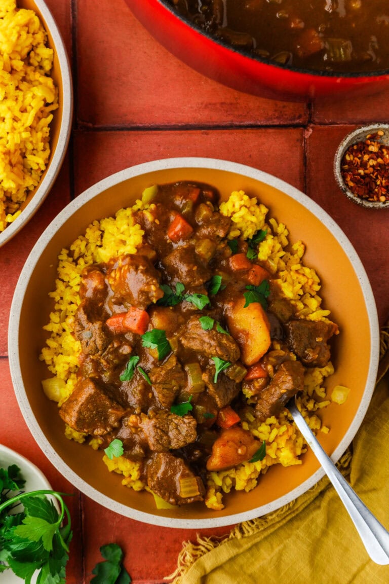 A bowl of yellow rice topped with beef stew containing chunks of meat, carrots, and potatoes, garnished with parsley. A spoon rests in the bowl, and side dishes with rice and herbs are visible nearby.