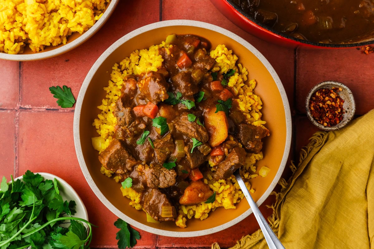 A bowl of yellow rice topped with hearty beef stew containing carrots, potatoes, and herbs. Nearby are bowls of yellow rice, parsley, red pepper flakes, and a pot of stew on a red tile surface.