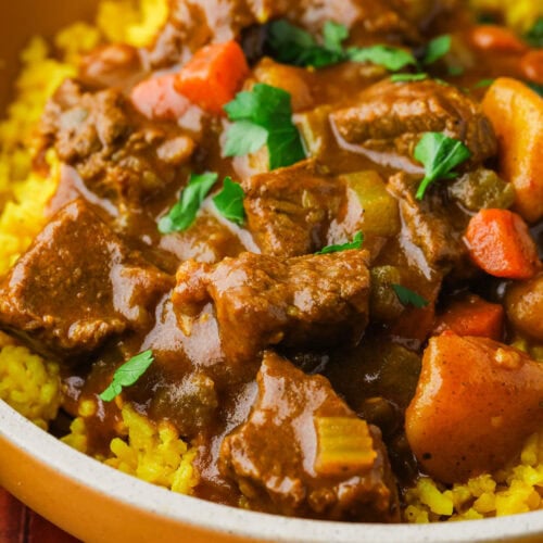 A close-up of a bowl filled with yellow rice, topped with beef stew containing chunks of beef, carrots, potatoes, and garnished with fresh parsley.