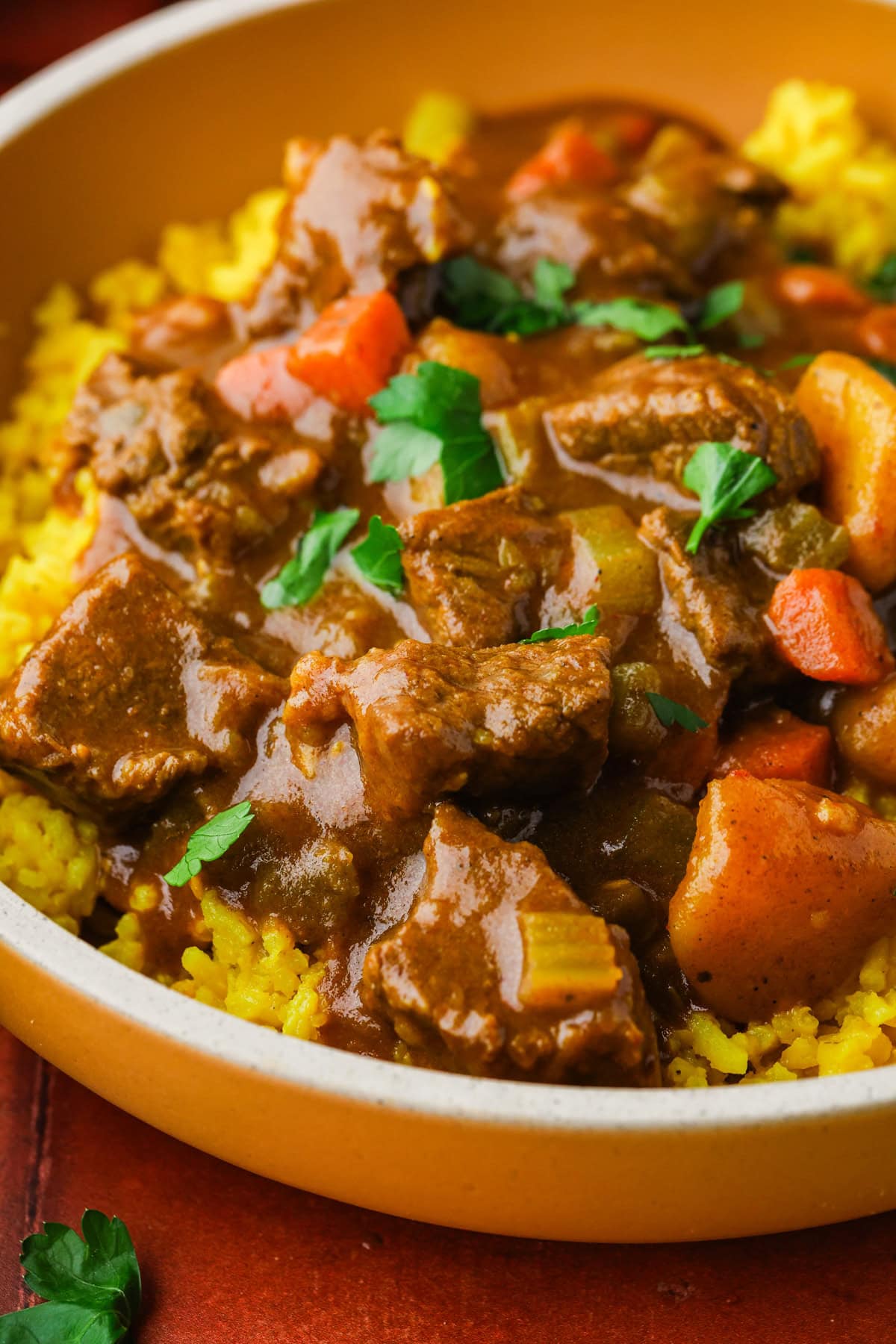 A close-up of a bowl filled with yellow rice, topped with beef stew containing chunks of beef, carrots, potatoes, and garnished with fresh parsley.