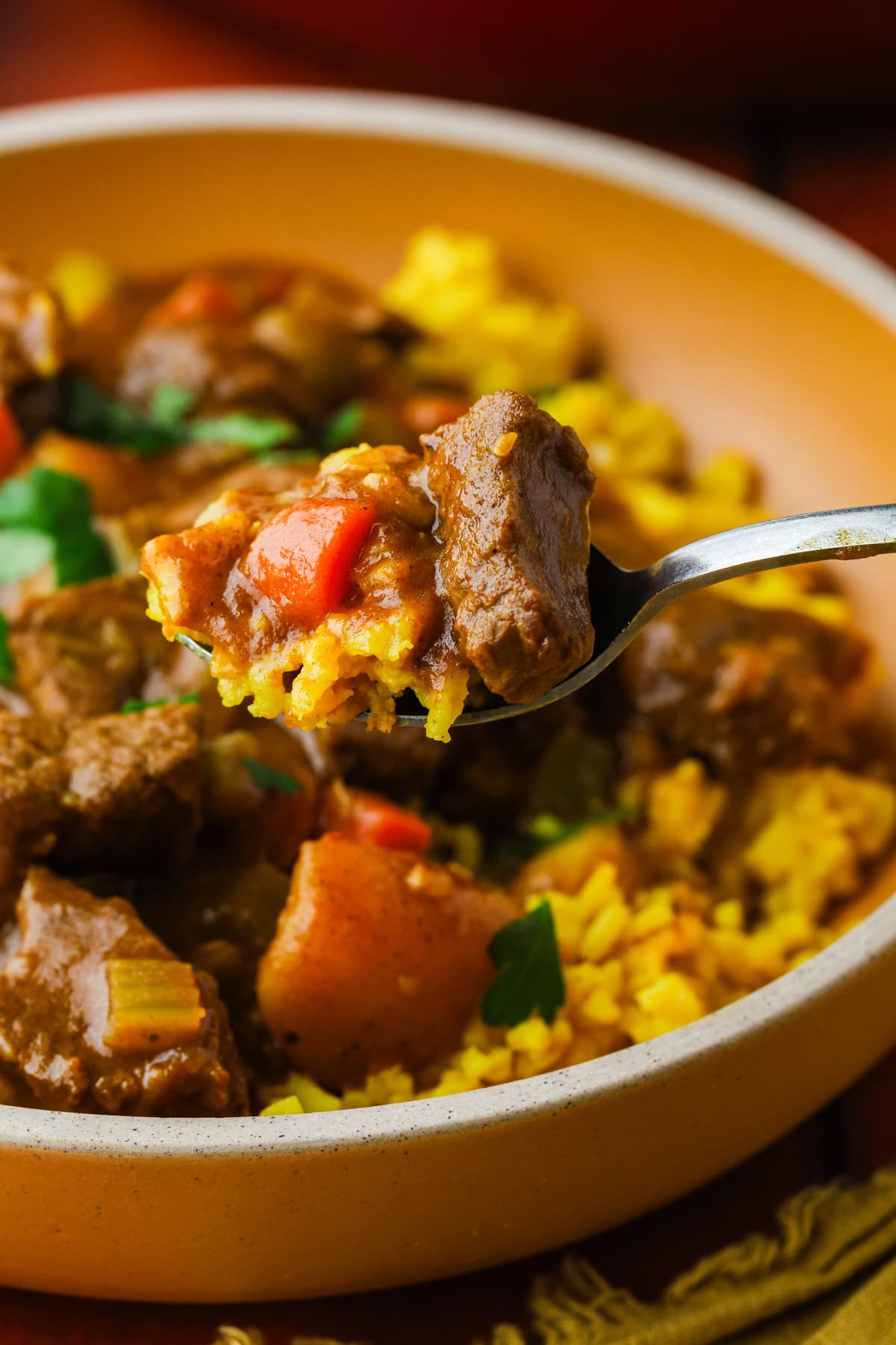 A close-up of a fork holding a bite of beef stew with chunks of beef, red bell pepper, and yellow rice, with more stew and rice in a bowl below.