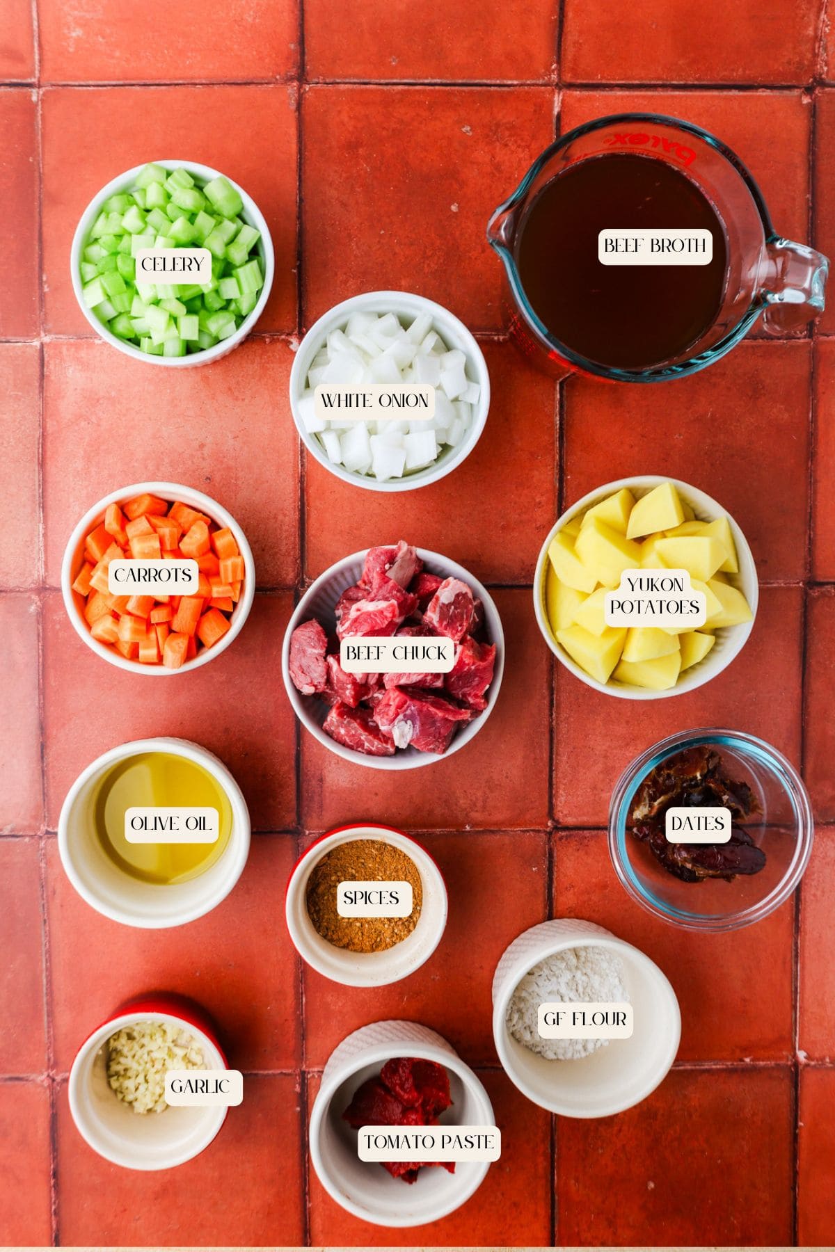 Top-down view of neatly arranged bowls containing labeled beef stew ingredients: celery, white onion, carrots, olive oil, garlic, beef chuck, spices, tomato paste, gluten-free flour, Yukon potatoes, beef broth, and dates on a red tiled surface.