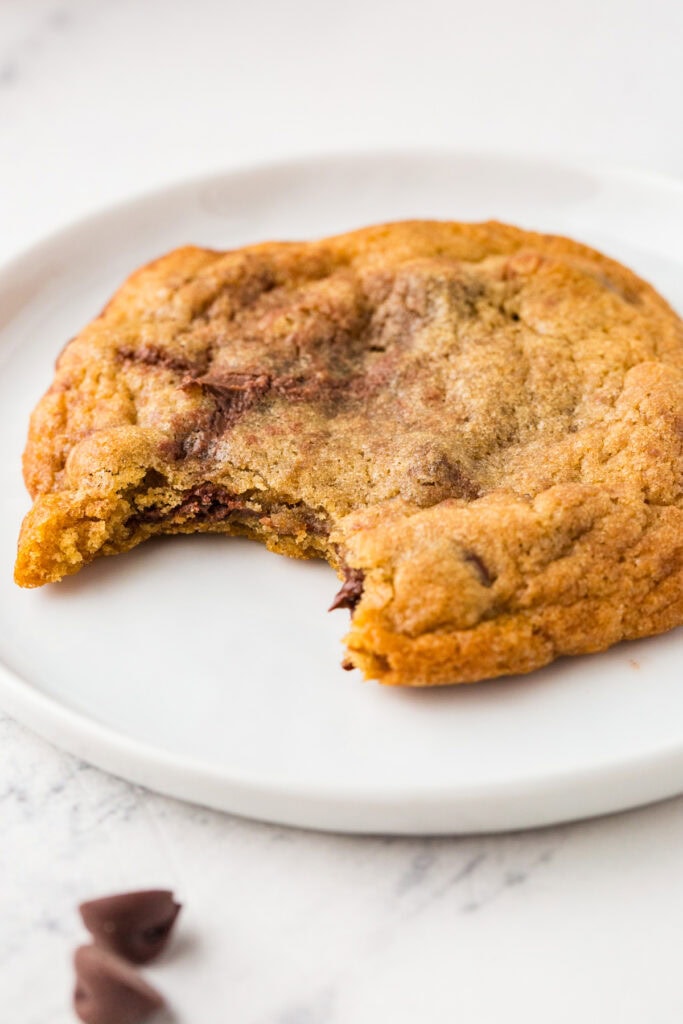 A chocolate chip cookie with a bite taken out sits on a white plate, perfect for showcasing in a Food Photography Portfolio. Nearby, a couple of chocolate chips rest on the pristine white surface.