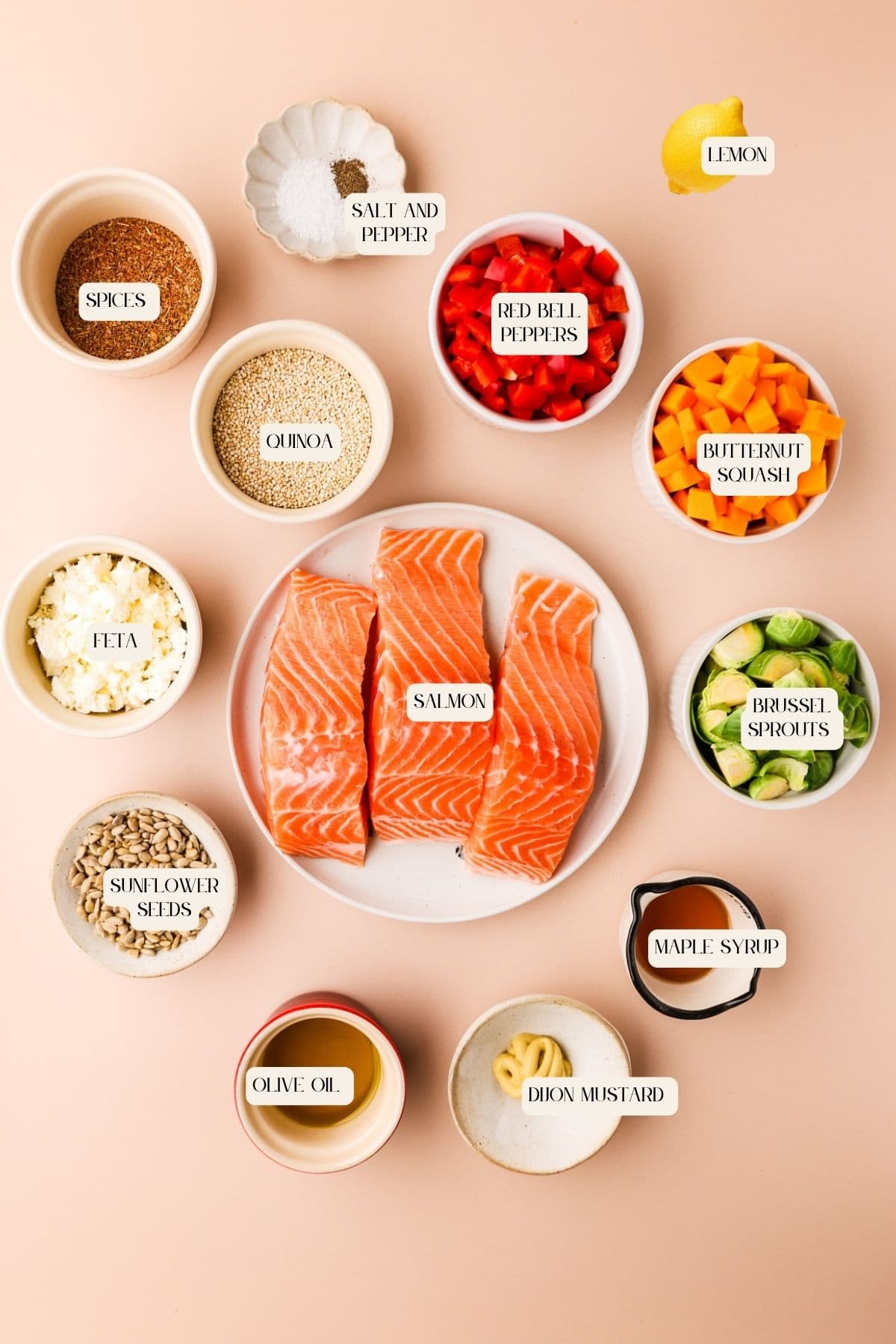 Top view of ingredients arranged on a light background: three raw salmon fillets in the center, surrounded by bowls of quinoa, feta, sunflower seeds, olive oil, maple syrup, Dijon mustard, spices, salt and pepper, red bell peppers, butternut squash, brussels sprouts, and a lemon wedge. Each item is labeled.