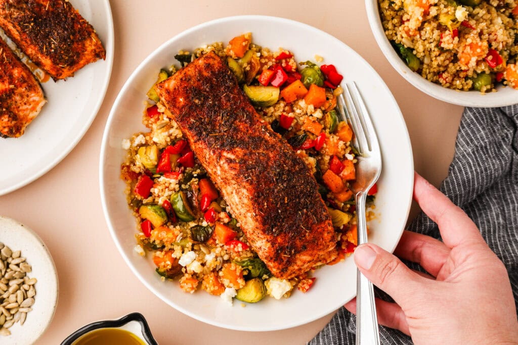 A hand holds a plate with a seasoned, cooked salmon fillet on top of roasted vegetables and couscous. A fork rests on the plate. Nearby are sunflower seeds, extra salmon, and a bowl of couscous.