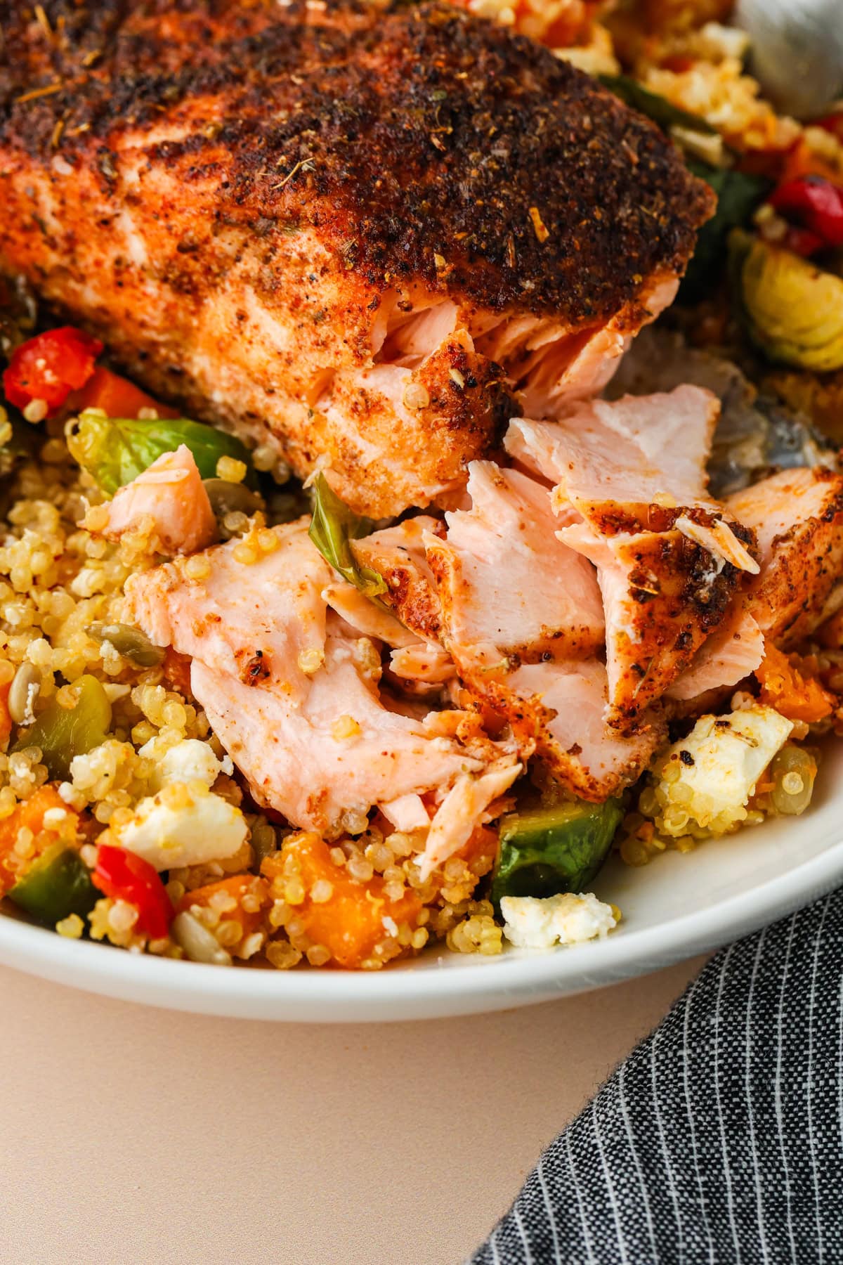 A close-up of a seasoned, cooked salmon fillet served on a bed of quinoa mixed with roasted vegetables and crumbled feta cheese on a white plate. A fork is flaking the salmon. A striped napkin rests nearby.