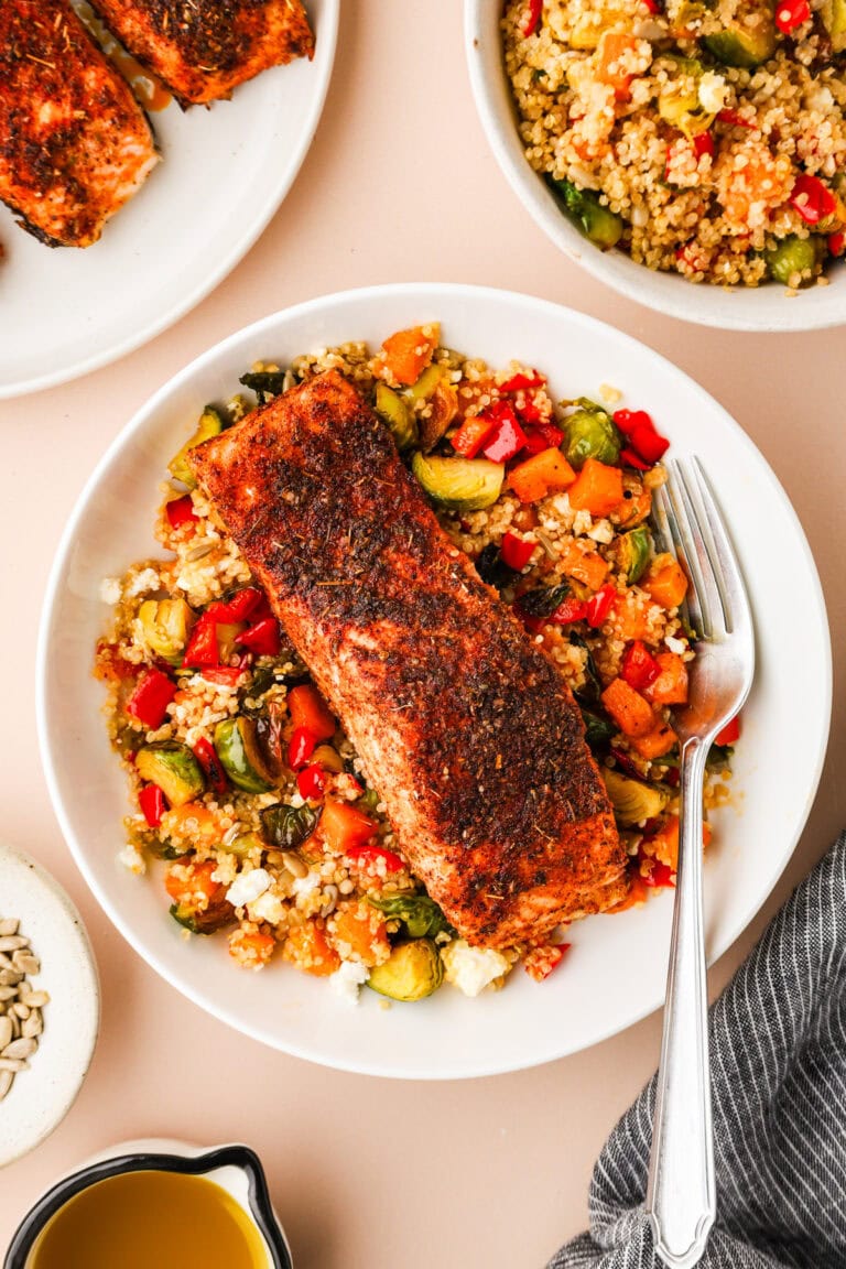 A plate with a seasoned, cooked salmon fillet on top of a colorful quinoa salad with red bell peppers, zucchini, and crumbled feta, beside a fork. Surrounding plates and a small bowl of dressing are partially visible.