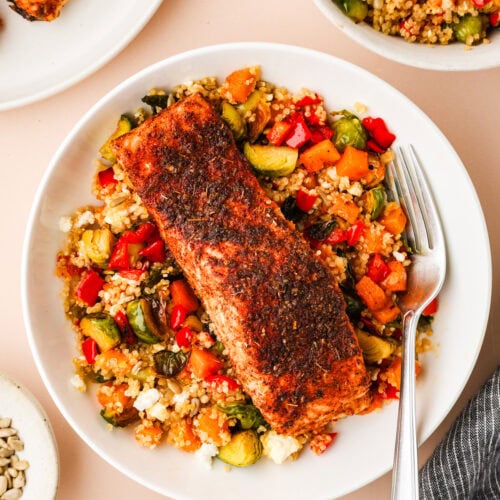 A plate with a blackened salmon fillet on top of couscous mixed with chopped vegetables and feta cheese, with a fork resting on the plate. Nearby are bowls of couscous, seeds, and dressing.