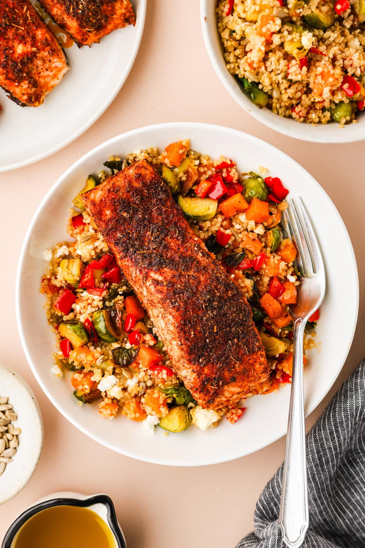 A plate with a blackened salmon fillet on top of couscous mixed with chopped vegetables and feta cheese, with a fork resting on the plate. Nearby are bowls of couscous, seeds, and dressing.